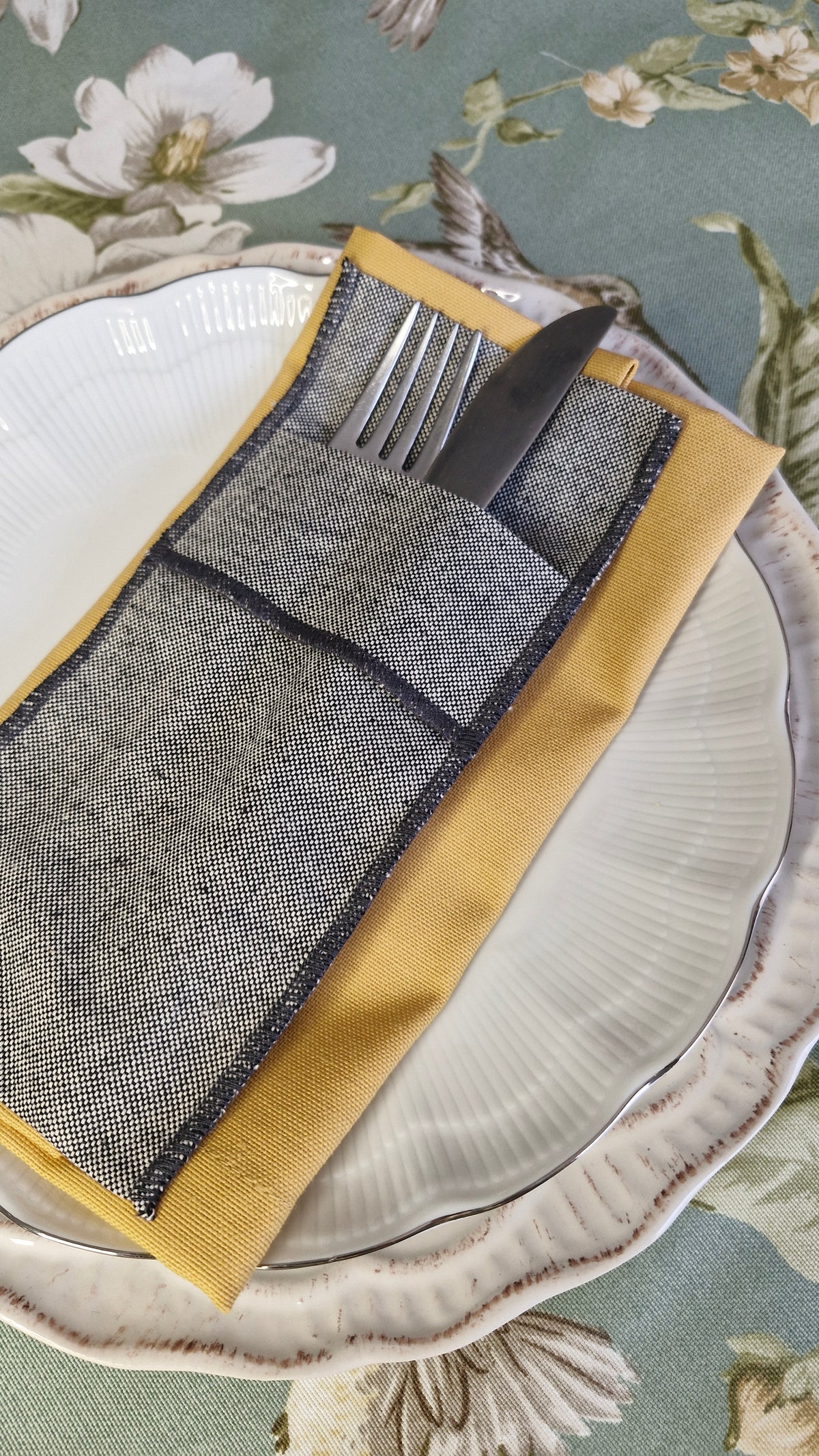 Servilleta mostaza sobre plato blanco con funda de cubiertos, en mesa con mantel verde floral